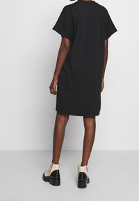 Personne portant une robe noire ample jusqu'aux genoux et des bottines beige et noires avec des clous, se tenant contre un fond gris uni.