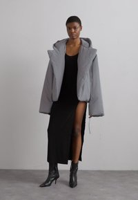 Šedá péřová bunda s oversized designem, zahrnující kapuci a široké rukávy. Nositá přes černé šaty s vysokým rozparkem a černými kotníkovými botami.