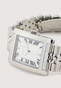 Sidabrinė nerūdijančio plieno laikrodis su stačiakampiu ciferblatu, baltu skaičiumi, juodais romėniškais skaičiais ir trimis dekoratyviniais deimantais. Laikrodžio apyrankė su grandinėlių dizainu.