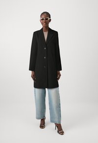 Cappotto nero su misura con revers e tre bottoni, abbinato a jeans a gamba larga blu chiaro e tacchi aperti argento.