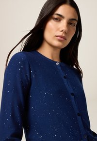 Frau mit langen dunklen Haaren, die eine marineblaue Strickjacke mit kleinen silbernen Pailletten und schwarzen Knöpfen vor einem neutralen Hintergrund trägt.