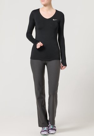 Kobieta ubrana w czarną, długorękawową bluzę Nike, ciemnoszare spodnie oraz fioletowe buty sportowe, stojąca z jedną zgiętą przed sobą ręką.