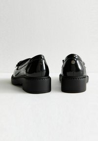 Chaussures en cuir verni noir avec un bout rond, un petit talon épais, une sangle décorative et des accents métalliques à l'arrière. Texture lisse et brillante.