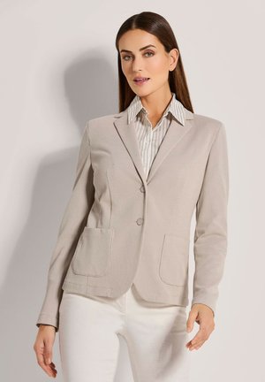 Blazer beige texturé avec revers crantés, deux poches à l'avant et une fermeture à un bouton, associé à une chemise à col rayée claire.