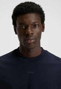 Mladý černoch s krátkými kudrnatými vlasy, oblečený v námořnickém modrém tričku s kulatým výstřihem a logem "HUGO" na přední straně, na čistém bílém pozadí.