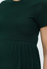 Σκούρο πράσινο ριμπ πλεκτό φόρεμα με κοντά μανίκια, εφαρμοστό μπούστο και ζώνη στη μέση σε άτομο με ανοιχτό χρώμα δέρματος.