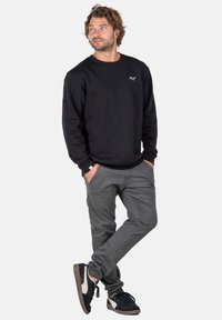 Mann mit welligem Haar, der einen schwarzen Sweatshirt, graue Hose und schwarz-weiße Sneaker trägt, steht mit den Händen in den Taschen und schaut nach links.