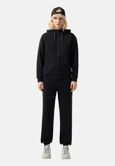 Ung person, der står og ser fremad iført sort hættetrøje med lynlås, matchende sorte joggers, hvide sneakers og sort kasket med bagsiden fremad på ensfarvet baggrund.