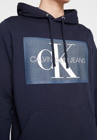 Tmavě modrá mikina s kapucí a šňůrkami, na přední straně velký potisk loga CK a text "CALVIN KLEIN JEANS". Měkký bavlněný materiál.