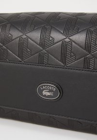 Sort Lacoste pung med strukturerede geometriske mønstre, glatte læderdetaljer og en sølv logo-plade.