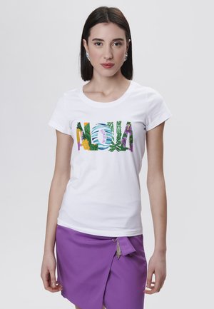 Hvid t-shirt med "ALOHA" grafik i flerfarvede tropiske mønstre. Korte ærmer og rund halsudskæring. Kombineret med en lilla nederdel.