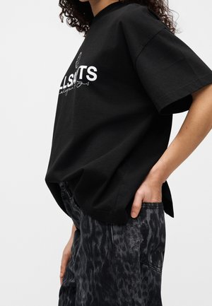 Le mannequin porte un T-shirt noir oversize à motif graphique et un pantalon foncé à motifs, avec une main glissée dans une poche.
