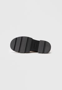 Semelle de chaussure en caoutchouc noir avec une surface texturée, comportant trois rainures horizontales pour une meilleure adhérence et une section de talon arrondie.