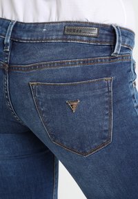 Vaqueros de mezclilla azul con un ajuste de tiro medio, que presentan un bolsillo trasero con un logo triangular plateado y detalles de costura en contraste.