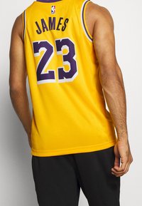 Hombre vistiendo una camiseta de baloncesto amarilla con el nombre "James" y el número "23" en púrpura, combinado con pantalones negros, visto desde atrás.