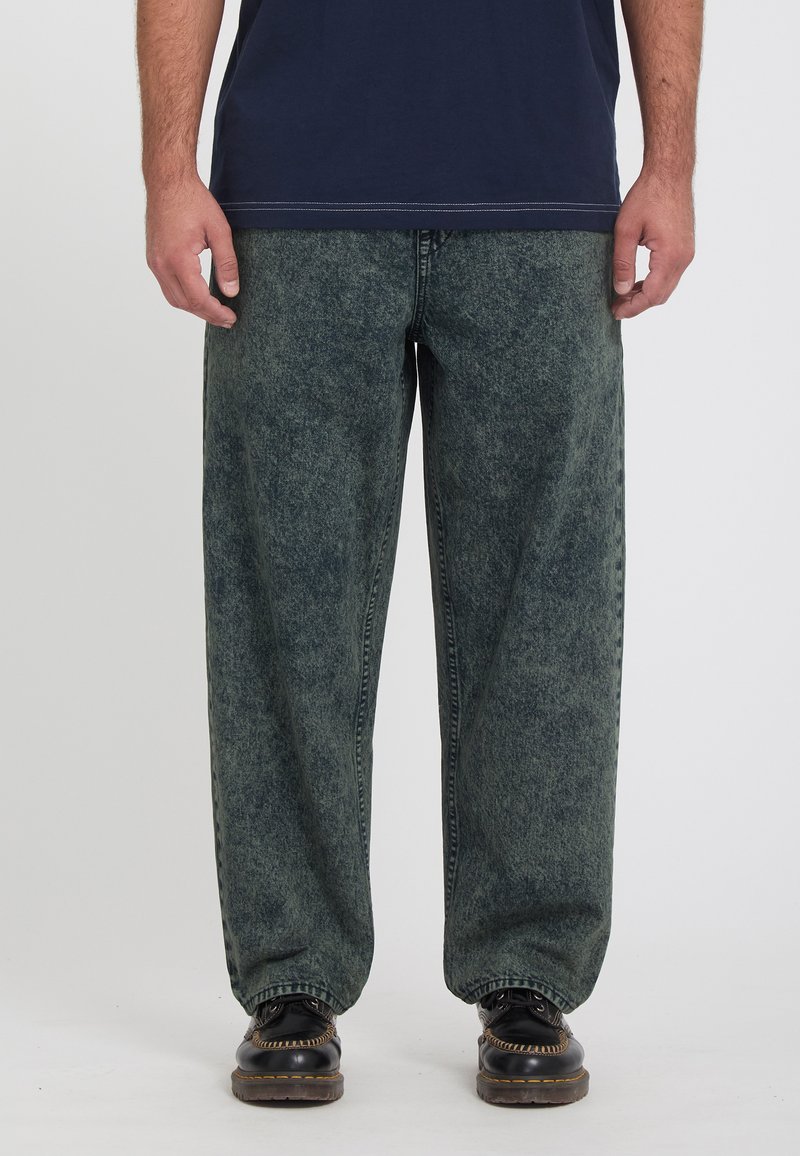 Άνδρας που φοράει φαρδιά τζιν με όξινο πλύσιμο, μαύρα δερμάτινα παπούτσια με κίτρινη ραφή και μπλε σκούρο πουκάμισο, στέκεται μπροστά σε απλό φόντο.