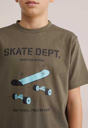 Dreng iført olivengrøn t-shirt med illustration af skateboarddele og tekst "SKATE DEPT. Lavet til bevægelse. Bygget til at rulle stykke for stykke."