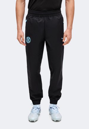 Bărbat purtând pantaloni sport negri cu logo albastru pe coapsă și crampoane albe, stând în picioare pe un fundal simplu și deschis la culoare.