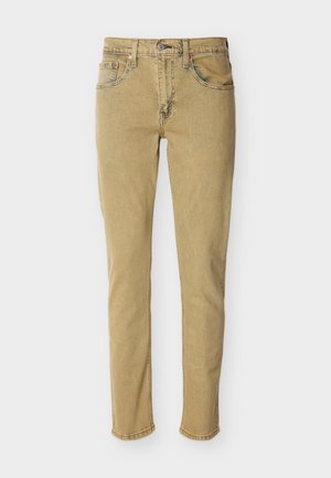 Béžové úzke rifle z denimu s prednými vreckami, zapínaním na gombík a zips, položené na rovnej ploche na bielom pozadí.