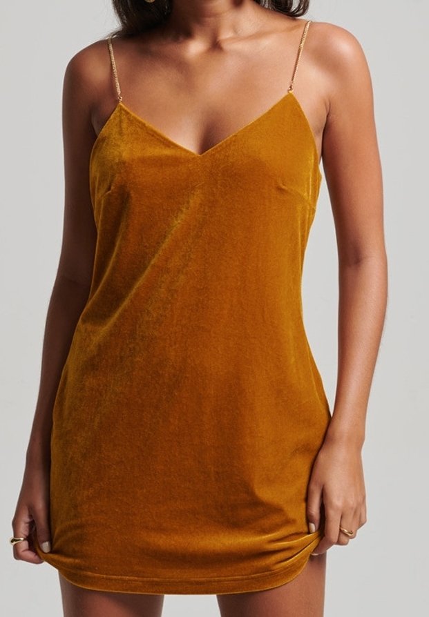 Femme portant une robe nuisette en velours jaune moutarde avec des bretelles fines en chaîne dorée, tenant légèrement l'ourlet des deux mains.