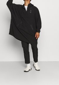 Schwarze Windjacke mit Kängurutasche, teilweisem Reißverschluss und elastischen Bündchen; kombiniert mit schwarzen Hosen und weißen Sportschuhen.