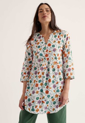 Femme debout, portant une longue tunique fleurie aux motifs orange, violet et vert, manches trois-quarts, et un pantalon vert sur fond uni.