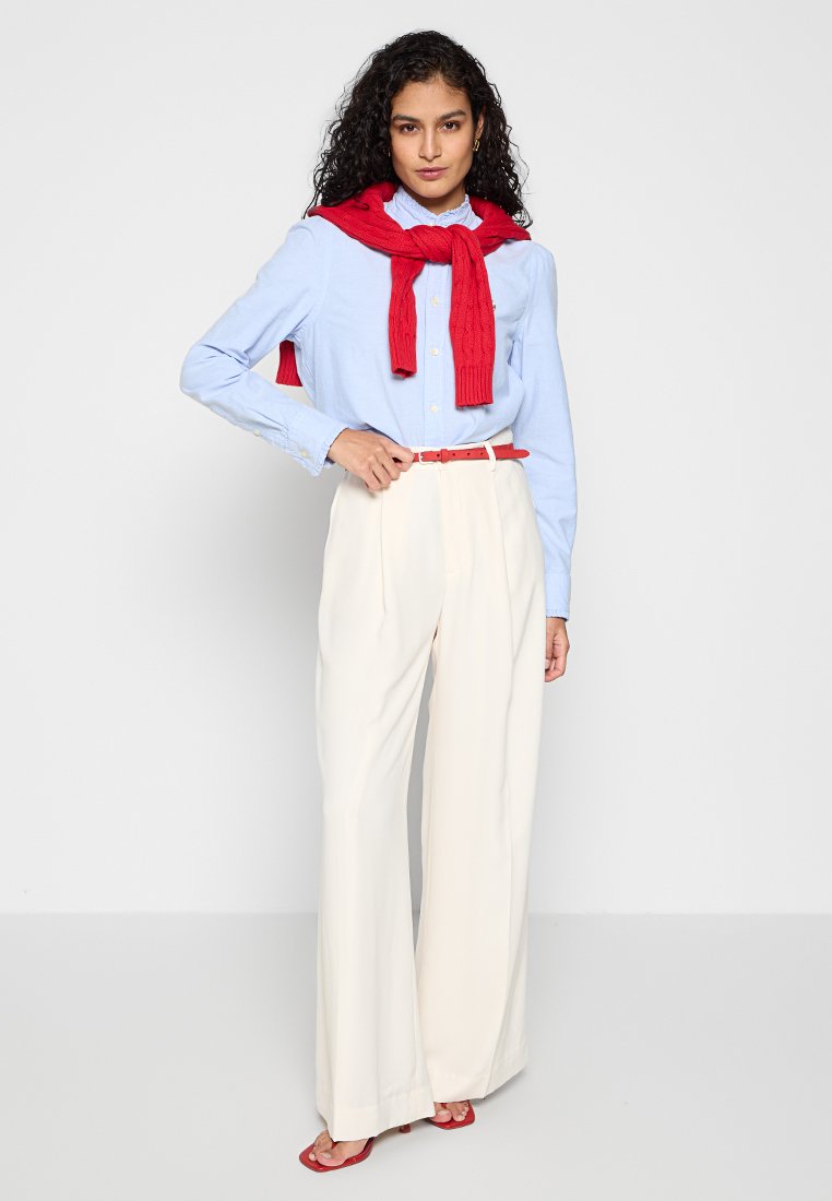 Helles blaues Hemd mit Knöpfen, roter Strickpullover um die Schultern gebunden, cremefarbene weite Hose, roter Gürtel und Sandalen, vor einem schlichten Hintergrund präsentiert.