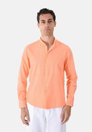 Uomo in piedi che indossa una camicia pesca a maniche lunghe abbottonata con colletto alla coreana e pantaloni bianchi su uno sfondo chiaro e uniforme.