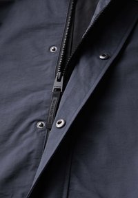 Chaqueta azul marino con una textura de tela suave, cremallera frontal completa, botones de metal y una etiqueta que dice "BUSCA AVENTURA."