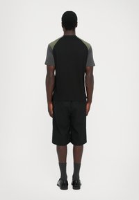 Man standing facing away wearing a color-blocked black, gray, and olive green t-shirt, black shorts, gray socks, and black shoes.