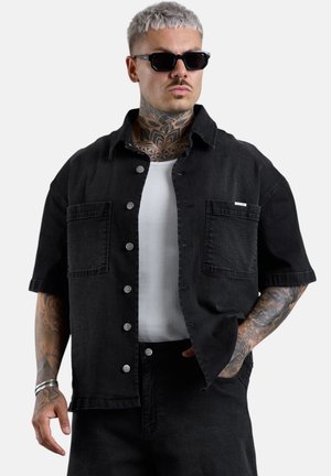 Mann mit hellgrauem Haar und Tattoos, der schwarze Sonnenbrille, ein schwarzes kurzärmeliges Hemd über einem weißen Top und schwarze Hose mit der Hand in der Tasche trägt.