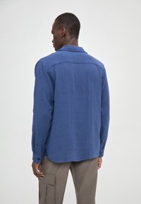 Cămașă albastră, texturată, cu mânecă lungă, guler și manșete cu nasturi, asortată cu pantaloni cargo de culoare deschisă; văzută din spate.