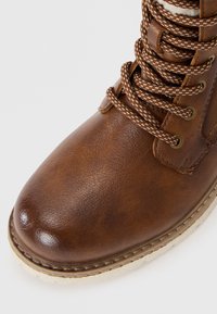 Botte en cuir marron avec surface texturée, bout rond et lacets à motifs marron et blanc. Elle est dotée d'une semelle en caoutchouc beige et d'œillets en métal.