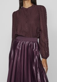 Blusa bordeaux con motivo a zigzag e chiusura a bottone, abbinata a una gonna plissettata, lucida, di un bordeaux più scuro con vita elasticizzata.