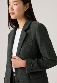 Tmavošedý herringbonový blazer s klopovým límcem, jednou kapsou a detailem na knoflíku. Hladká textura, strukturovaný tvar a jemné logo na rukávu.