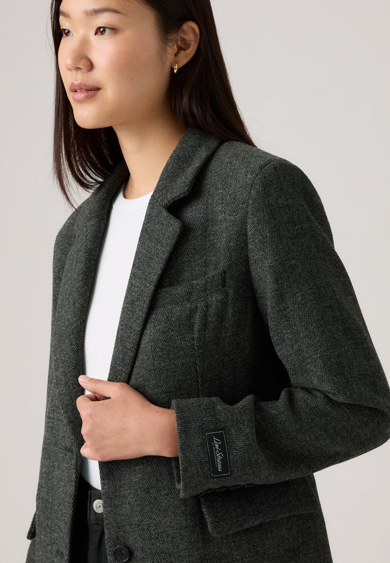 Mörkgrå herringbone-blazer med spårad krage, en ficka och knappdetalj. Jämn textur, strukturerad form och subtil varumärkeslogotyp på ärmen.