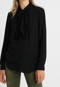 Svart långärmad blus med en slipsdetalj vid kragen, med en slät textur och en avslappnad passform.