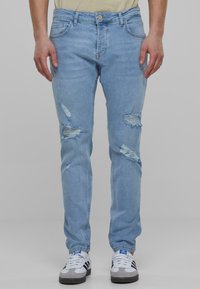 Unos jeans de mezclilla azul claro presentan múltiples parches desgastados, un corte slim, estilo de cinco bolsillos y un cierre de botón en la cintura.