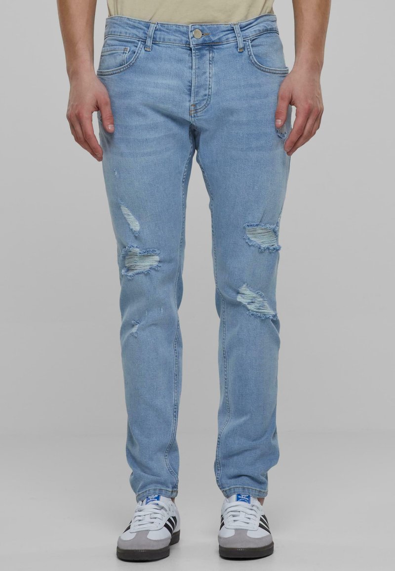 Unos jeans de mezclilla azul claro presentan múltiples parches desgastados, un corte slim, estilo de cinco bolsillos y un cierre de botón en la cintura.