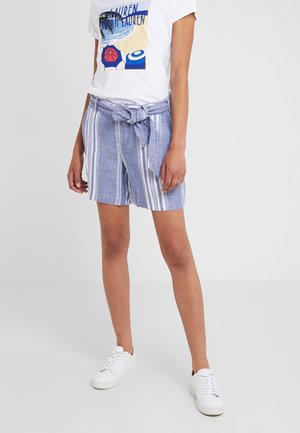 Persona que lleva una camiseta blanca con diseño de playa, pantalones cortos de talle alto a rayas azules y blancas con cinturón anudado y zapatillas blancas de pie.
