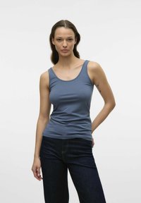 Débardeur bleu en tissu extensible, avec un décolleté en forme de creux et une coupe ajustée. Associé à un jean en denim foncé. Texture lisse.
