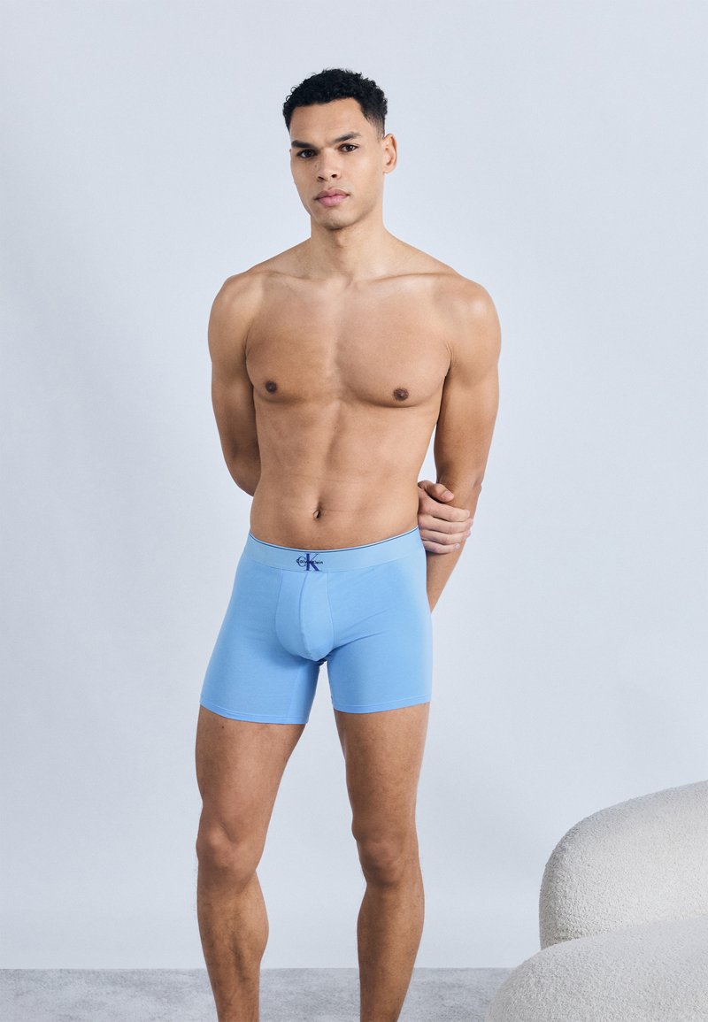 Άνδρας σε φόρμα που στέκεται με τα χέρια πίσω από την πλάτη, φοράει ανοιχτό μπλε μποξέρ, με φόντο απλό ανοιχτό γκρι.