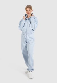 Felpa azzurra chiara con zip frontale e cappuccio oversize, abbinata a pantaloni jogger coordinati con vita elastica e polsini.