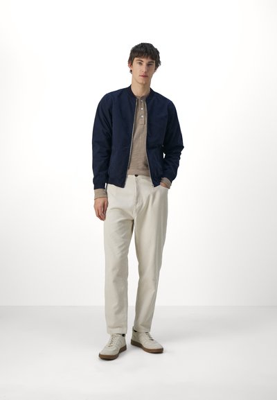 Chaqueta de piloto navy con cremallera, camiseta henley color beige, pantalones crema con textura, y zapatillas blancas con suelas beige. Diseño sencillo.