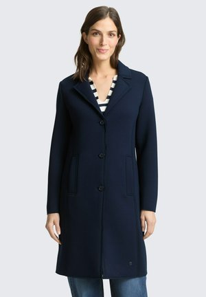 Kobieta ubrana w granatowy, długi płaszcz na paskowaną bluzkę i niebieskie dżinsy, stojąca na tle jednolitego, jasnego tła.