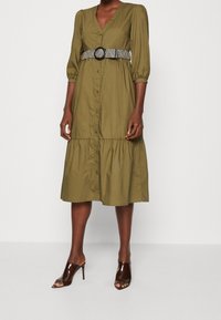 Oliwkowa midi sukienka z dekoltem w serek, zapinana na guziki, z marszczonymi rękawami. Posiada wzorzysty czarno-biały pasek i warstwowy dół. Brązowe klapki z odkrytymi palcami.