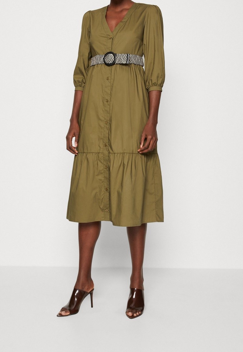 Oliwkowa midi sukienka z dekoltem w serek, zapinana na guziki, z marszczonymi rękawami. Posiada wzorzysty czarno-biały pasek i warstwowy dół. Brązowe klapki z odkrytymi palcami.