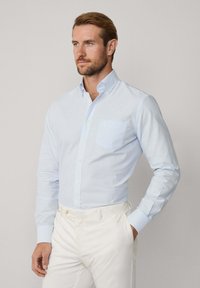 Lichtblauwe gestreepte blouse met lange mouwen, een enkele borstzak en witte broek. Glad materiaal met een getailleerde pasvorm.