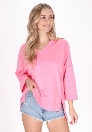 Rosa V-Ausschnitt-Oberteil mit Dreiviertelärmeln, aus weichem Stoff gefertigt. Kombiniert mit blauen Denim-Shorts, verfügt es über einen lockeren Schnitt und seitliche Schlitze.