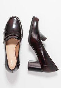 Chaussures à talon en cuir bordeaux foncé avec un bout rond, une sangle décorative et un talon bloc. Texture lisse avec des détails de couture contrastants.